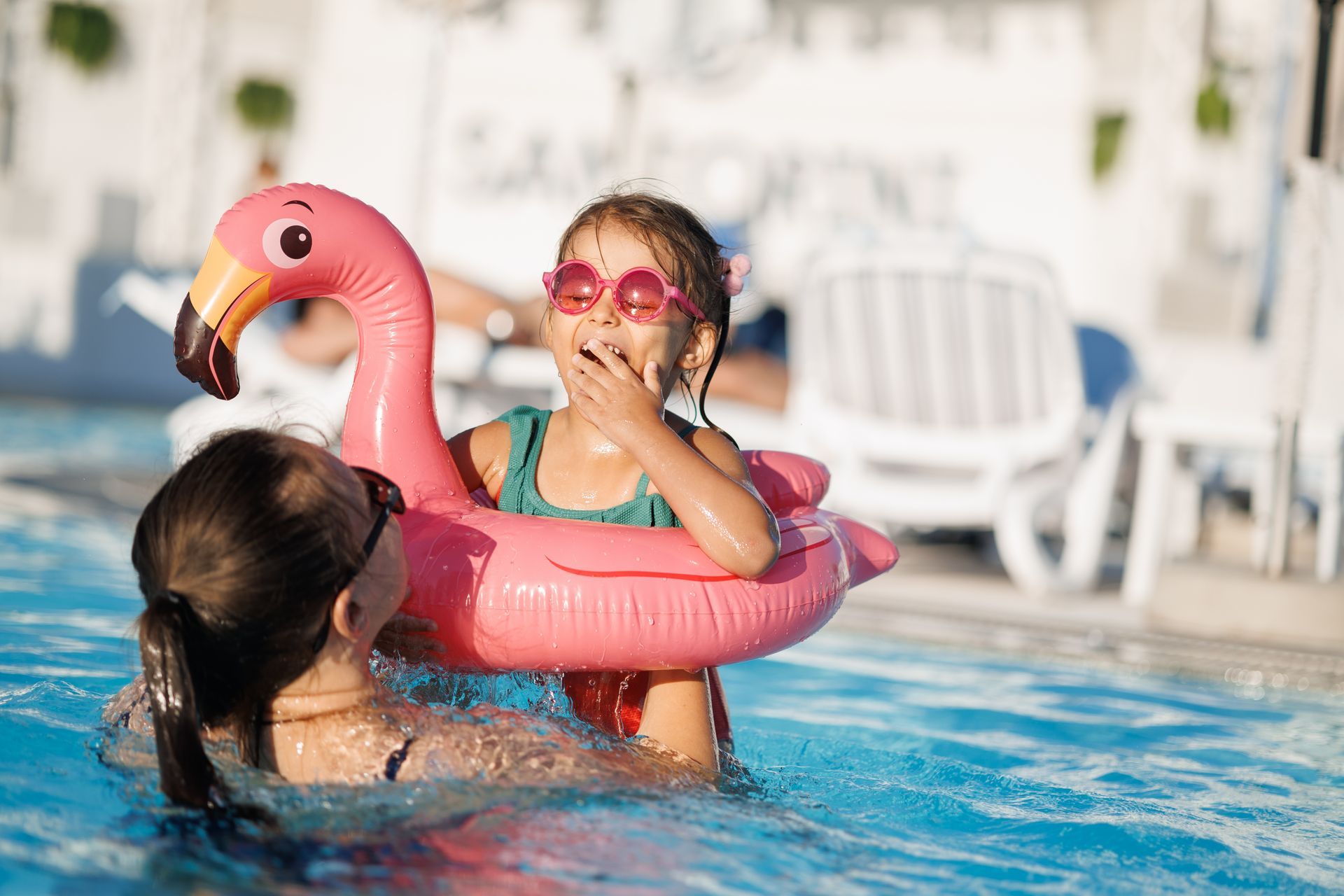 Two children in pool: one in a pink flamingo float, wearing sunglasses, looking surprised. Other child holds onto the float.
