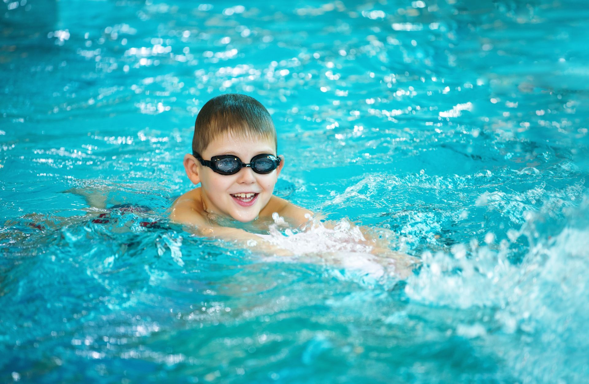 Boy swimming in a blue pool, wearing goggles and smiling.
