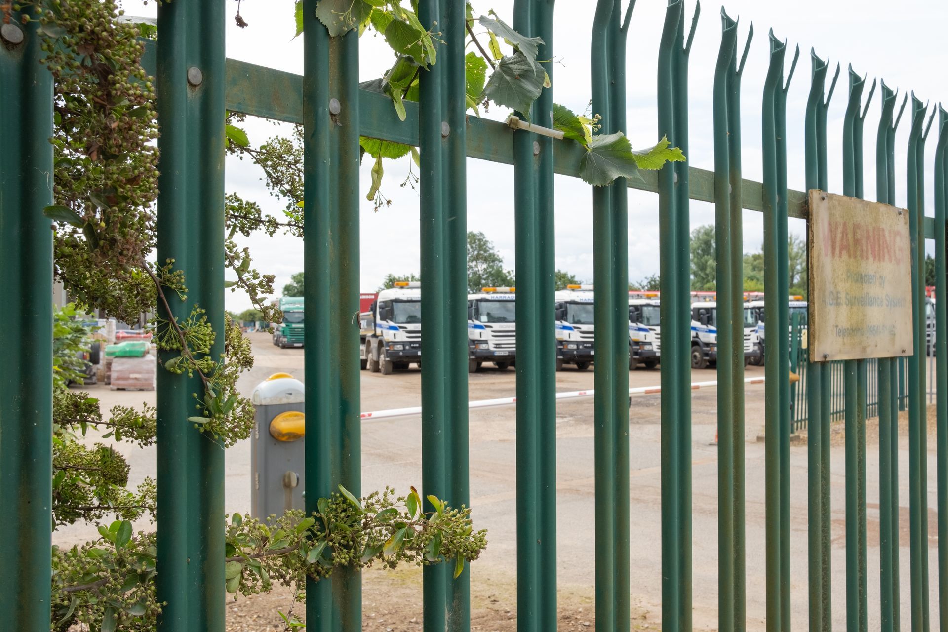 A green palisade fence with a warning sign guards a yard of fleet trucks. A green palisade fence with a warning sign guards a yard of fleet trucks.