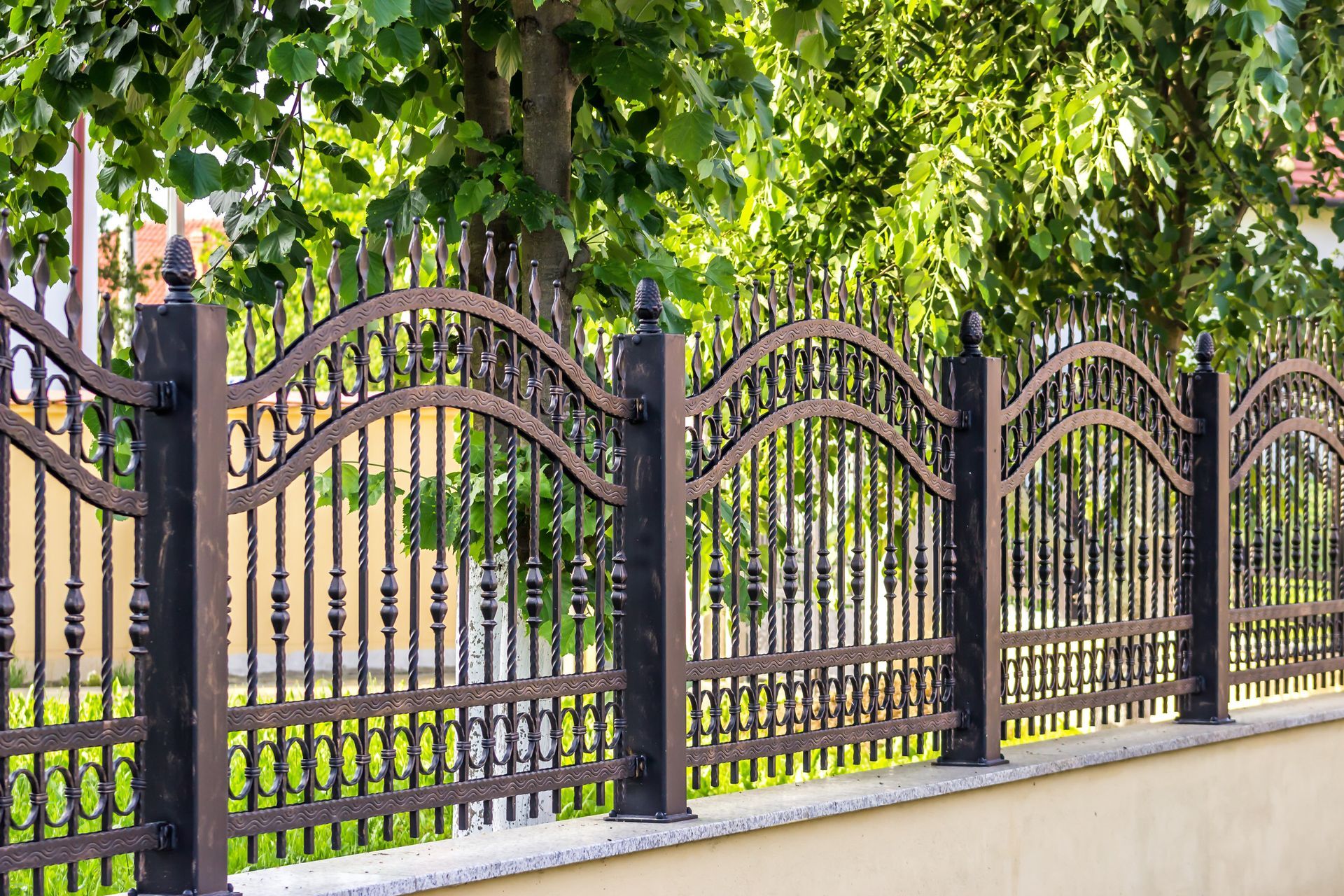 Black wrought iron fence with decorative arches, set on a beige concrete wall, with trees in the background.