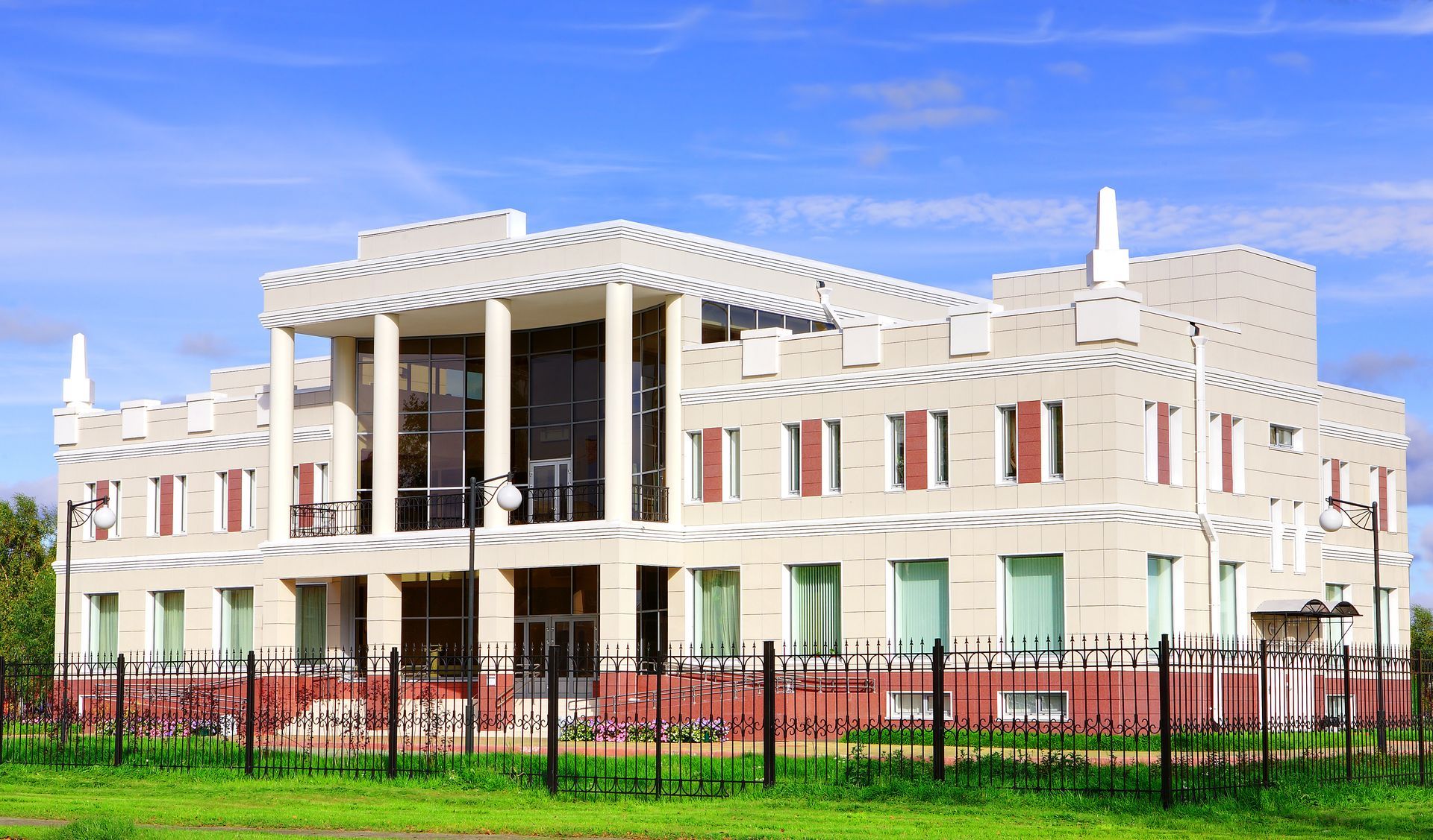 Two-story building with white walls, red accents, and a crenelated roof, behind a black fence on a grassy lot under a blue sky.