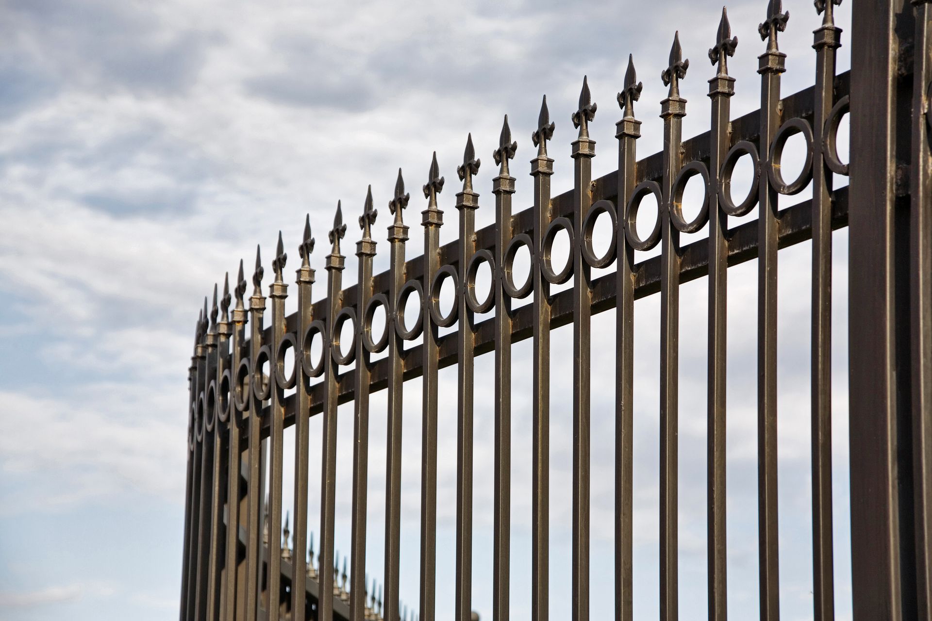 Curved metal fence with spikes and cloudy sky in the background.