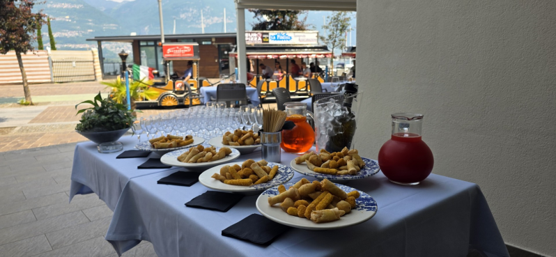In un ristorante, su un tavolo apparecchiato con una tovaglia bianca all'aperto, sono disposti piatti di antipasti fritti e una caraffa di bevanda rossa.