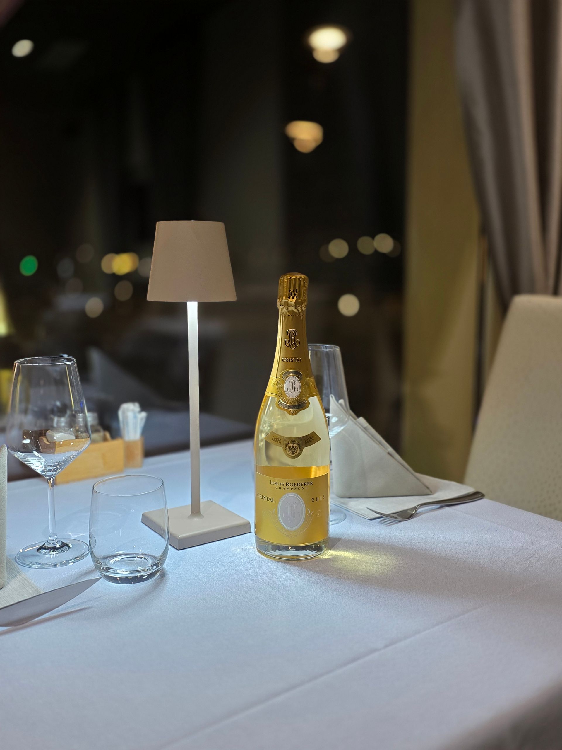Un tavolo con una tovaglia bianca, una piccola lampada, una bottiglia di champagne e due bicchieri in un ristorante scarsamente illuminato di sera.