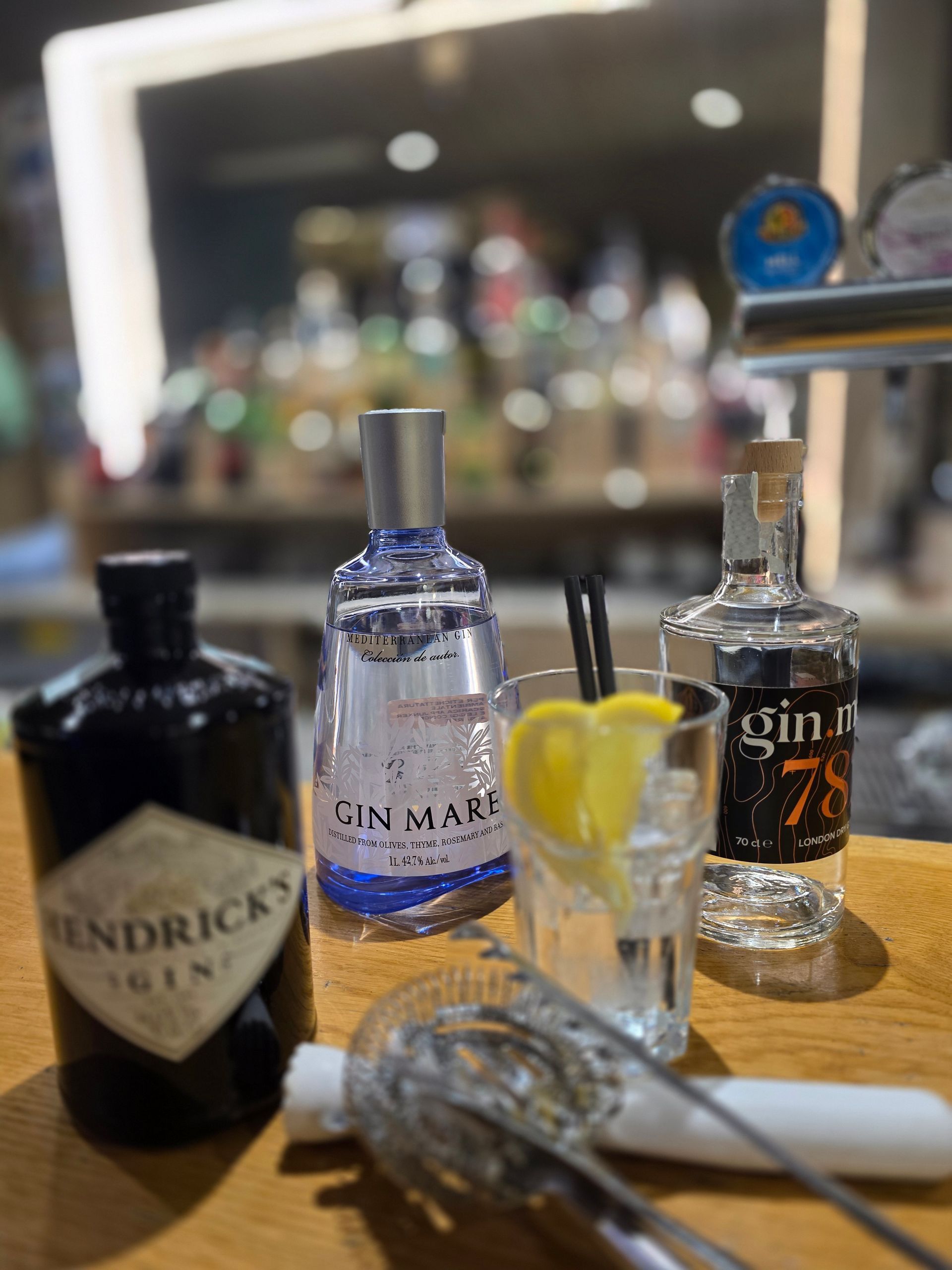 Tre bottiglie di gin e un bicchiere guarnito con utensili da bar su un bancone di legno, in uno sfondo sfocato e illuminato da una luce soffusa.