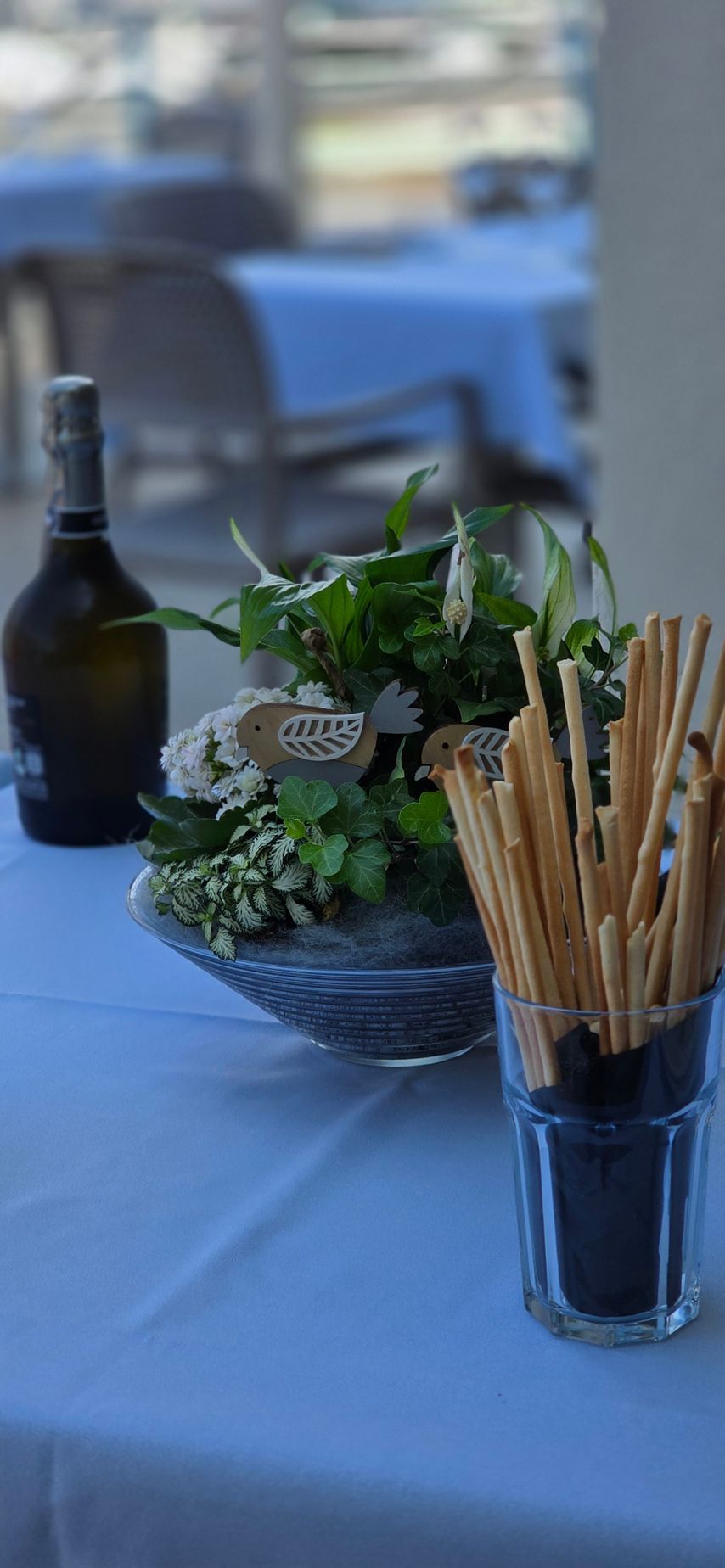 In un caffè all'aperto, su una tovaglia azzurra, una bottiglia di vino, un bicchiere di grissini e una piccola composizione floreale sono appoggiati su una bottiglia di vino, un bicchiere di grissini e un piccolo mazzo di fiori.