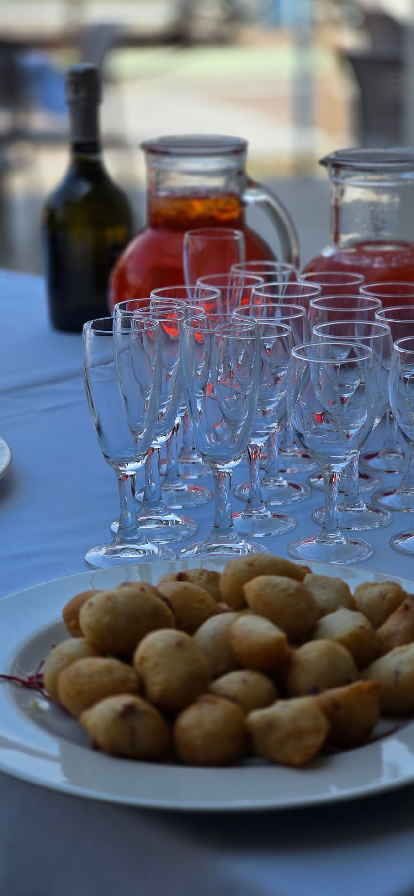 In primo piano si trova un piatto di snack fritti, con file di flûte di vetro vuote, due brocche e una bottiglia alle sue spalle.