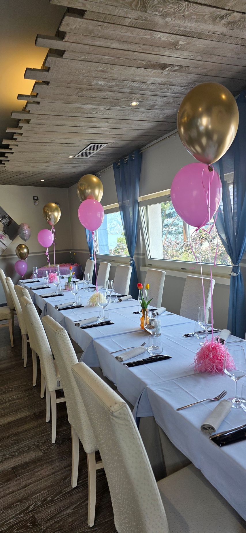 Un lungo tavolo apparecchiato per una festa in una stanza con un soffitto rustico, con centrotavola di palloncini rosa e oro.