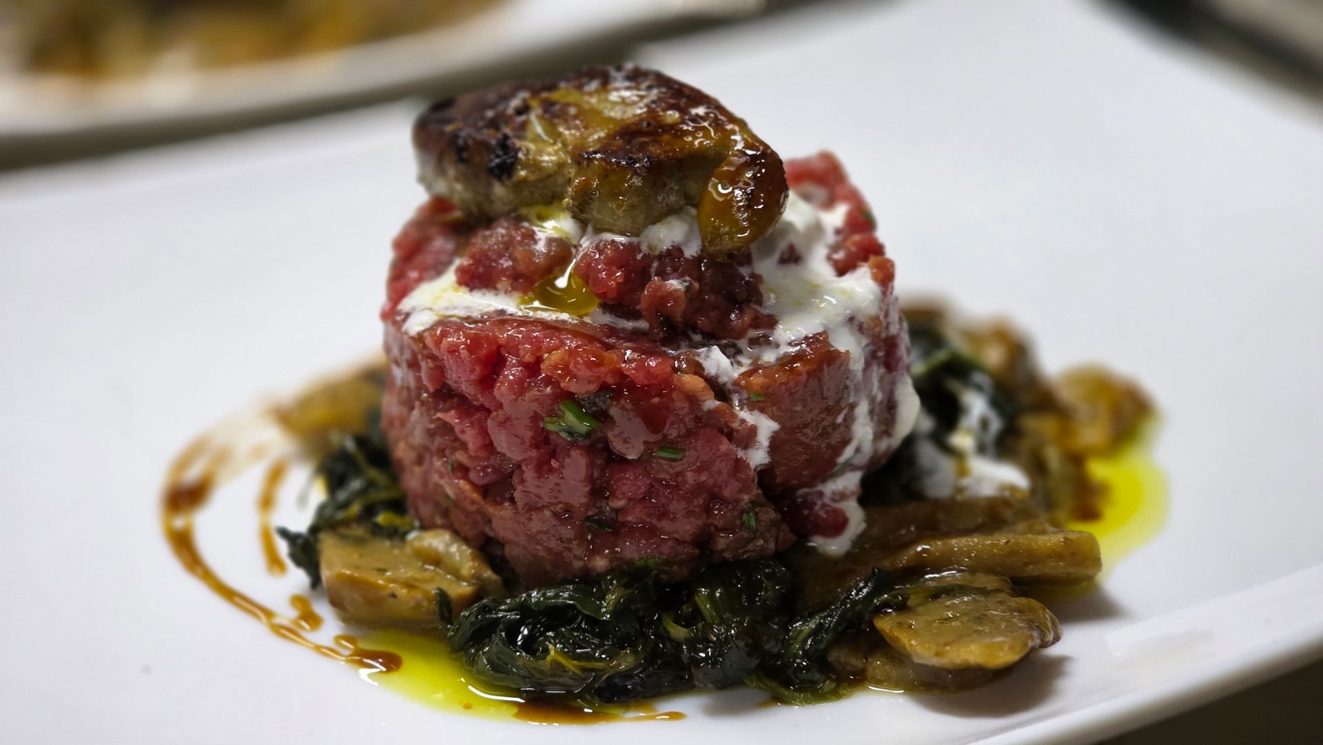 Un cilindro di tartare di manzo sormontato da foie gras scottato, guarnito con verdure e funghi saltati, servito su un piatto bianco.