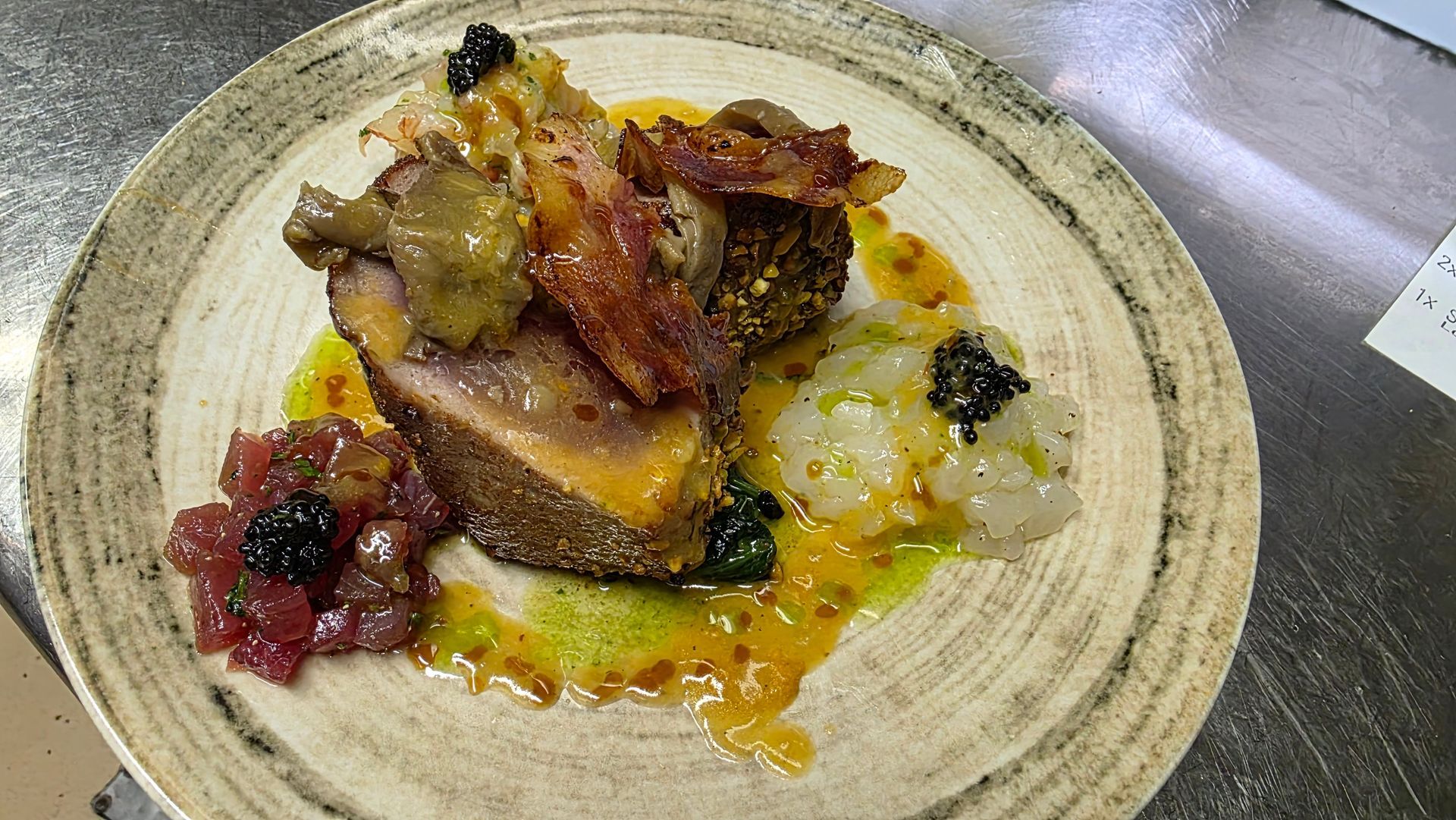 Un piatto con un cuore di carne scottata, guarnito con pancetta, accompagnato da tonno rosso tritato, cipolla a dadini e caviale.