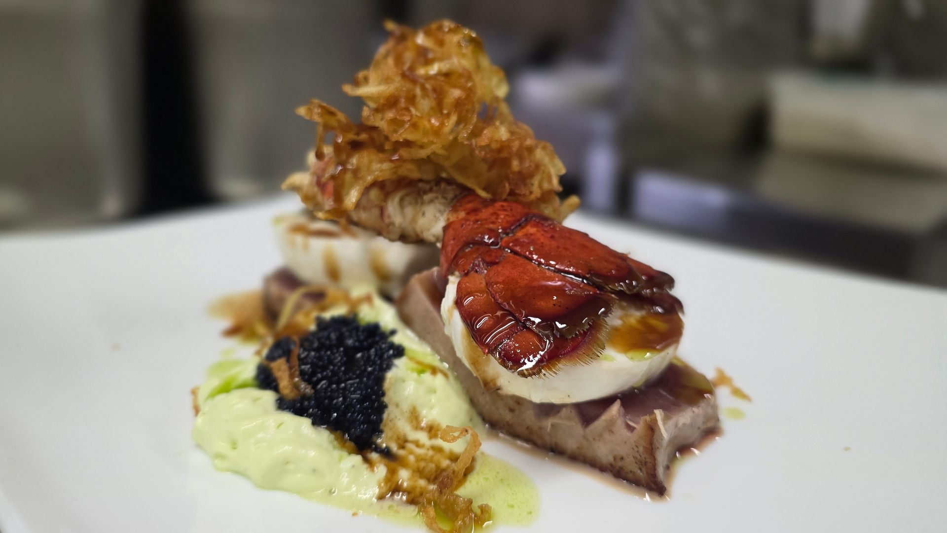 Un antipasto gourmet a base di tonno scottato, uovo in camicia, coda di aragosta, caviale e cipolle fritte su una leggera salsa verde.