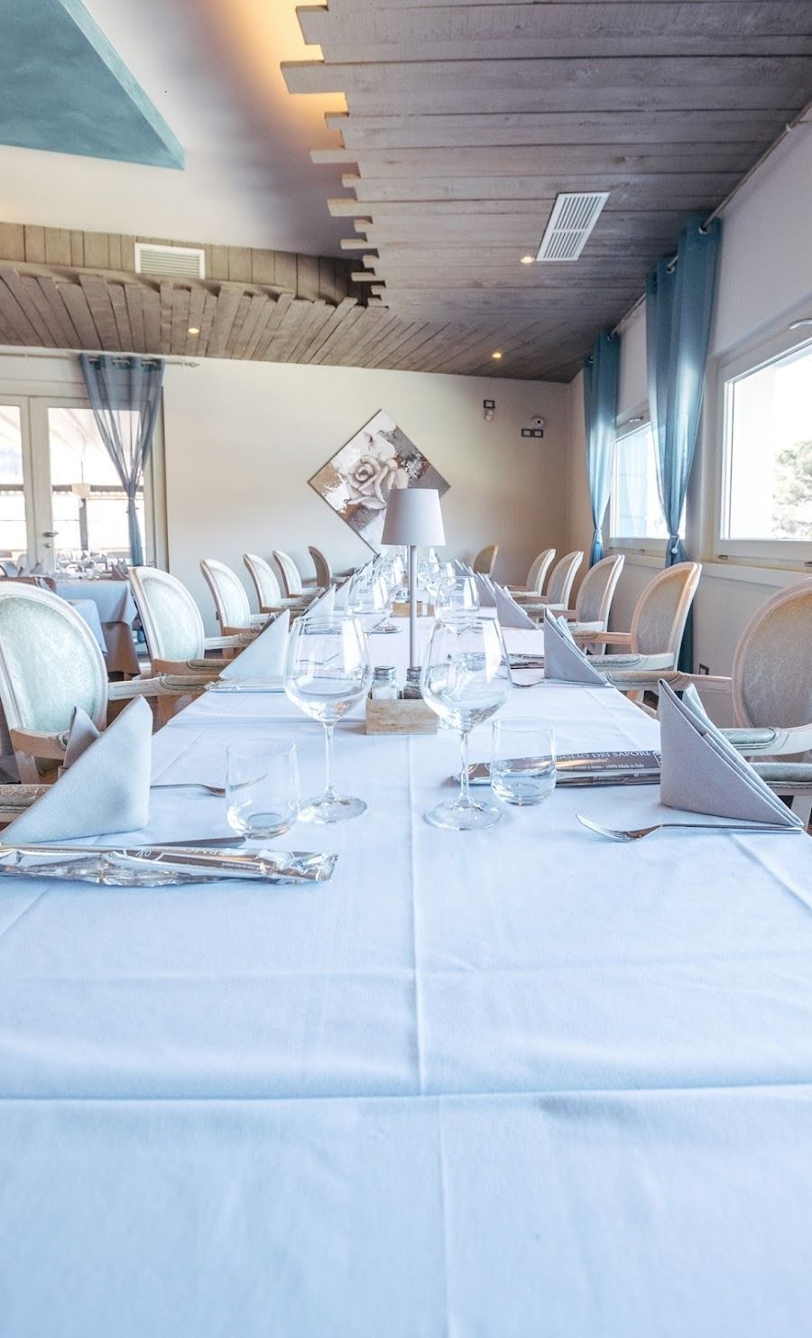 In un ristorante con tende blu, un lungo tavolo da pranzo formale apparecchiato con tovaglie, tovaglioli e bicchieri bianchi.