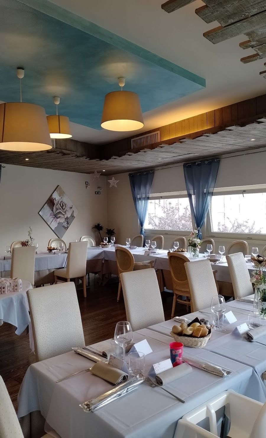 Una sala da pranzo allestita per un evento formale, con lunghi tavoli apparecchiati con tovaglie bianche, sedie beige e una delicata illuminazione a sospensione.