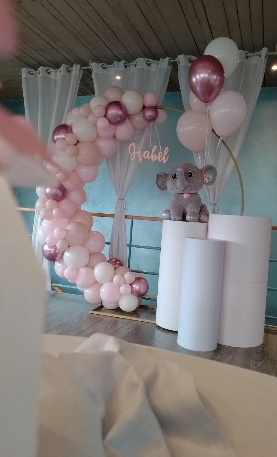 Un arco di palloncini a forma di mezzaluna nei toni del rosa e del bianco, con un elefantino di peluche in cima a piedistalli cilindrici bianchi.