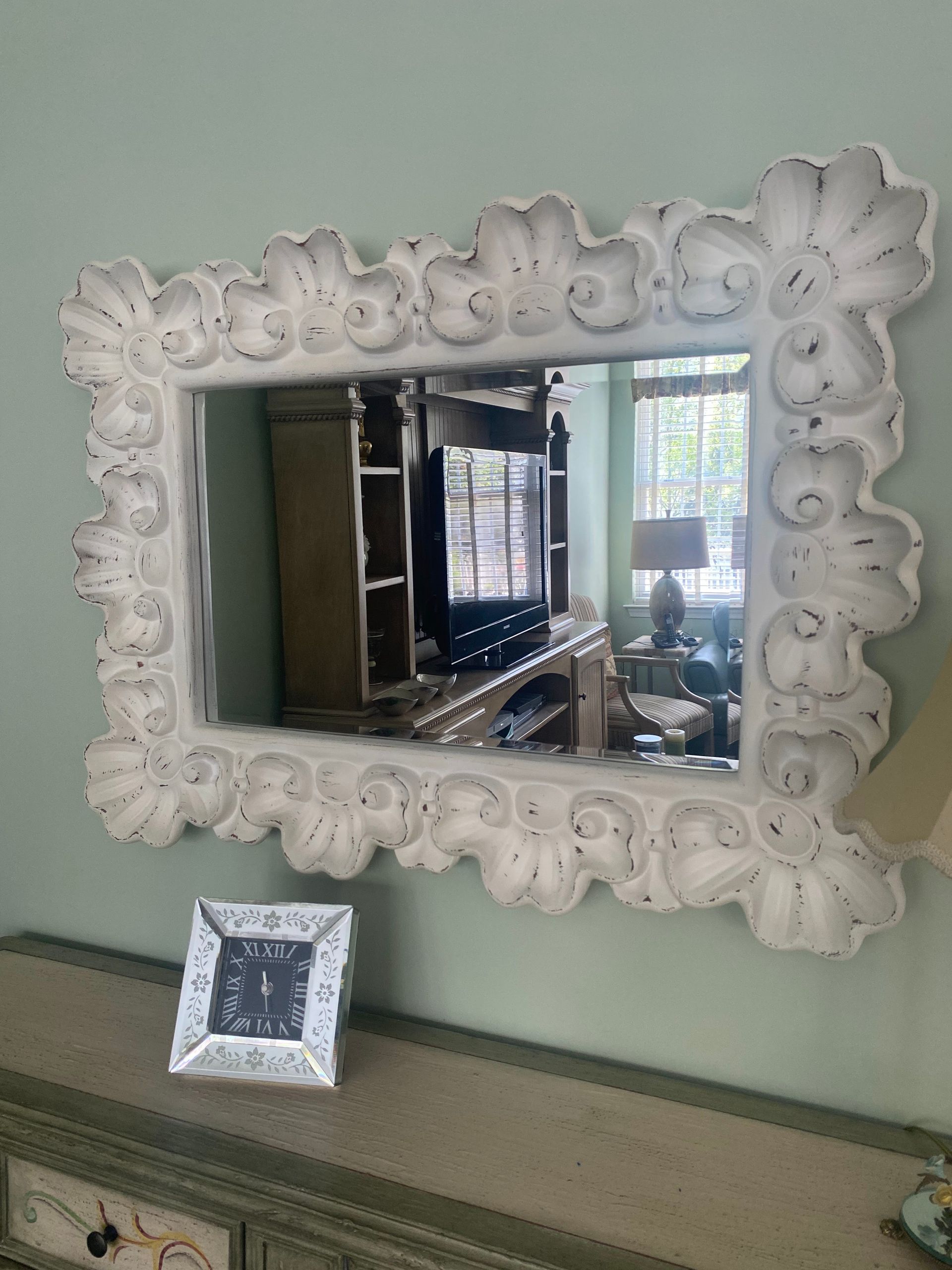 White, ornate rectangular mirror hanging on a light-green wall above a gray console table..