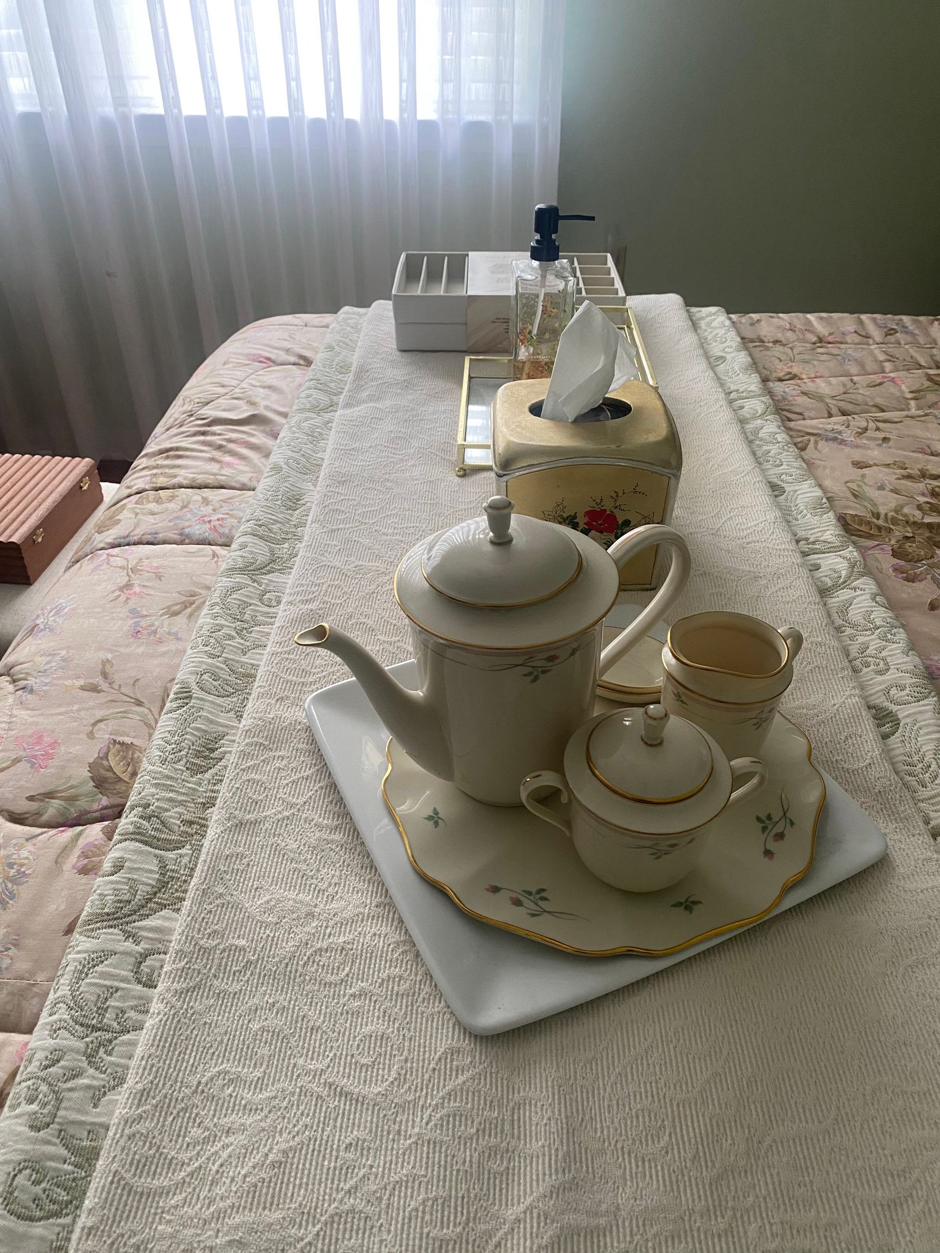 Tea set on a bed runner, with a tissue box, soap dispenser, and window in the background.