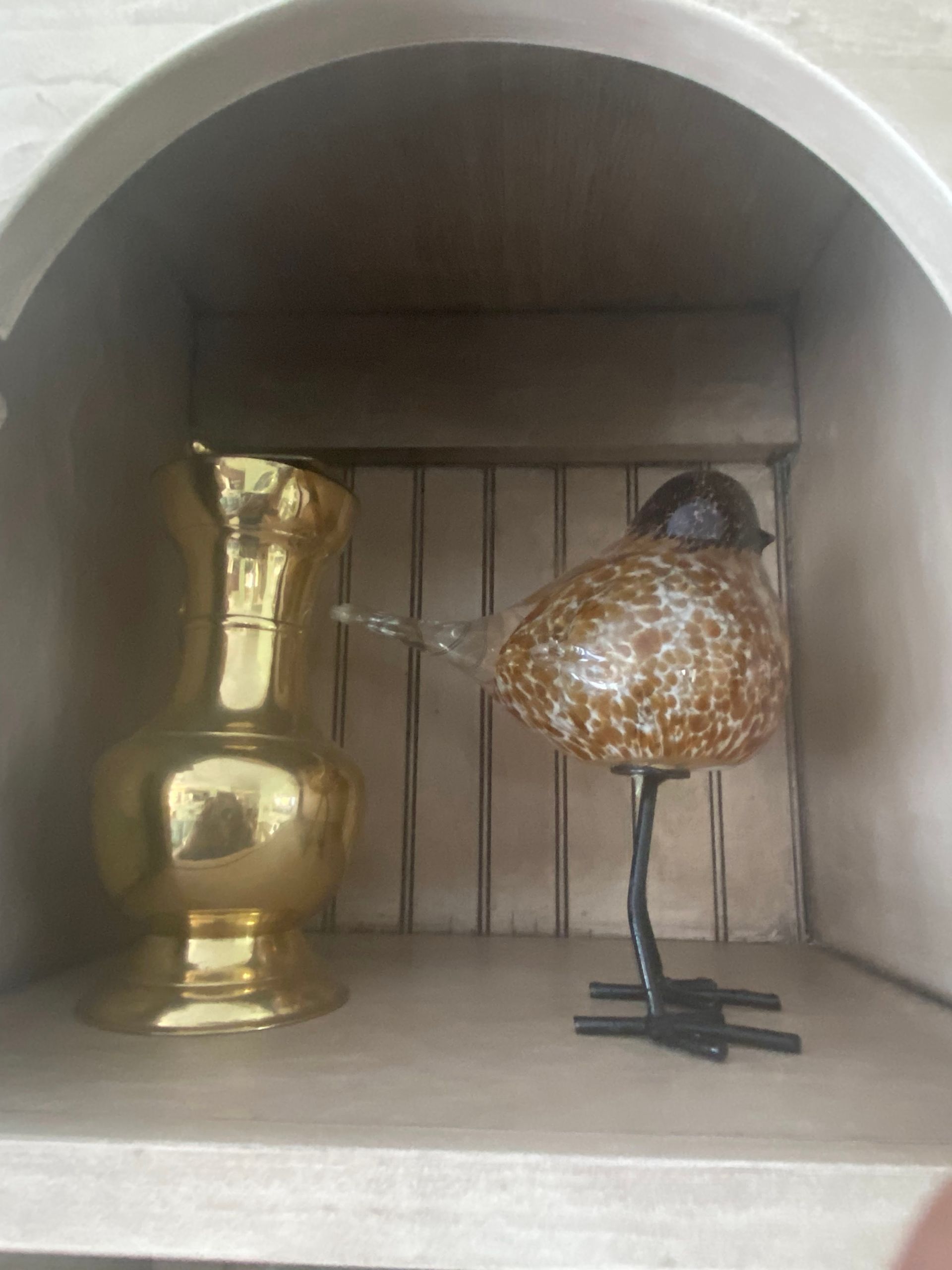 Gold pitcher and spotted glass bird on a metal stand displayed inside a recessed shelf.