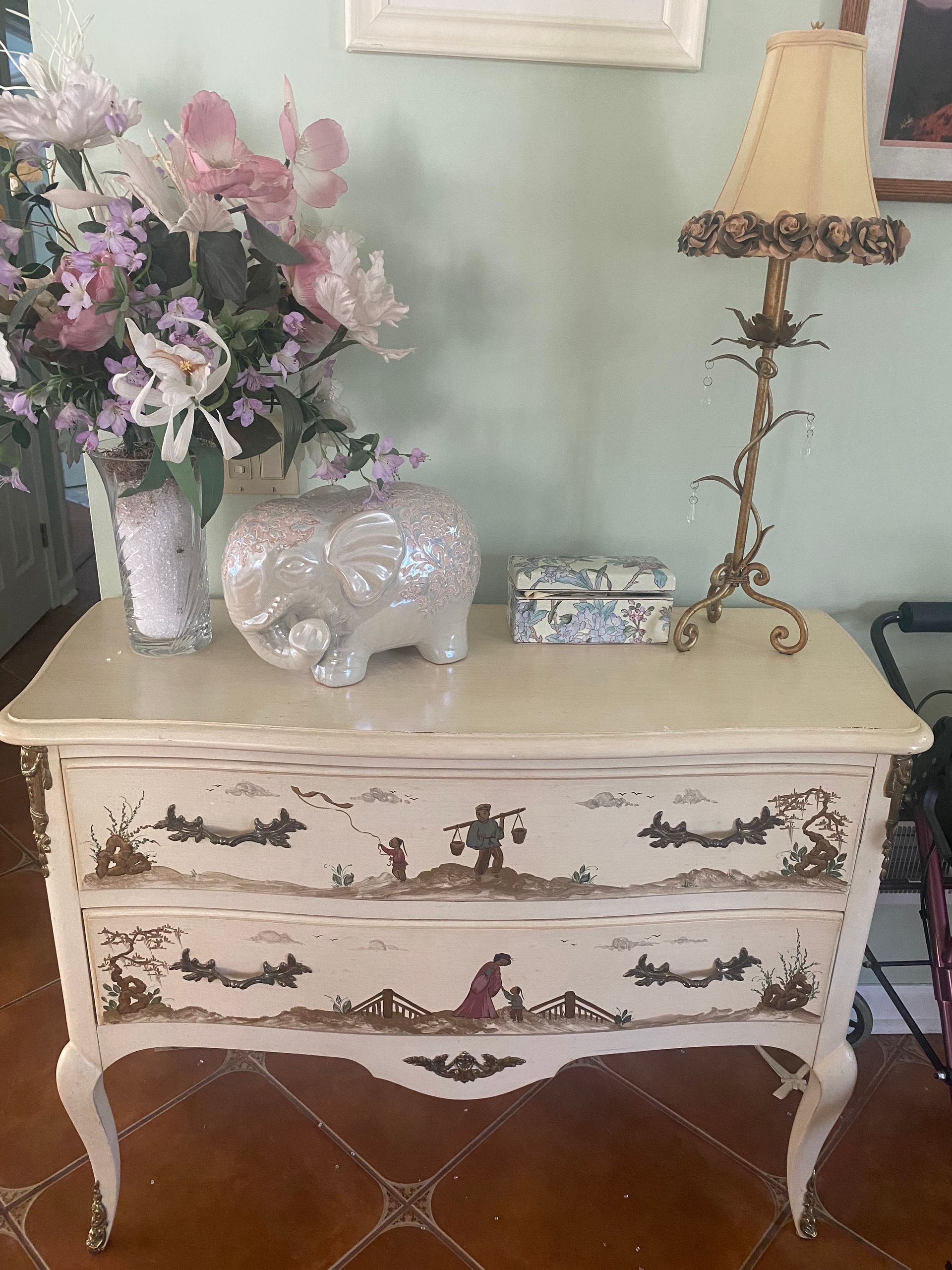 Cream-colored nightstand with landscape painted drawers, topped with flowers, a pig figurine, and a lamp.