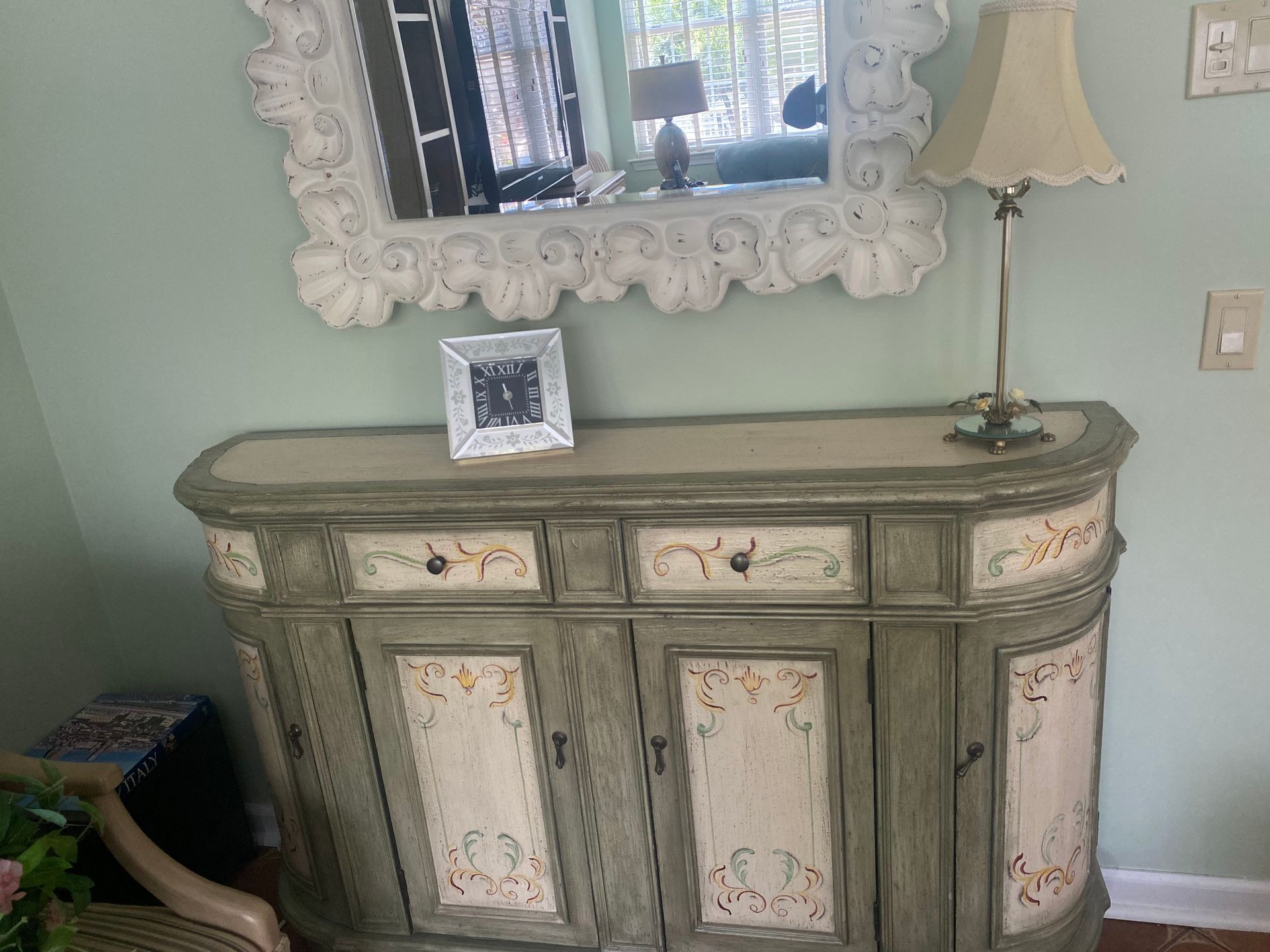 A green and cream painted cabinet with a decorative mirror and lamp against a green wall.