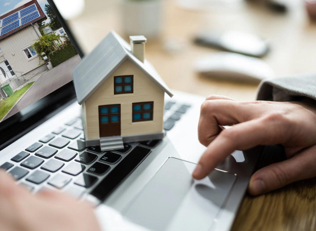 Person using a laptop, a small house model on the keyboard; a house image is on the screen.