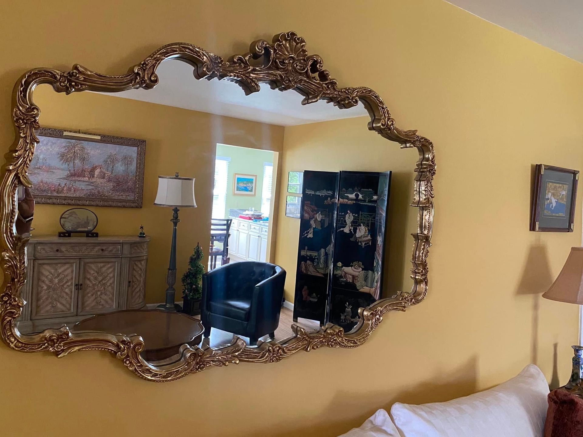 Ornate gold framed mirror on yellow wall, reflecting a room with furniture and an interior doorway.