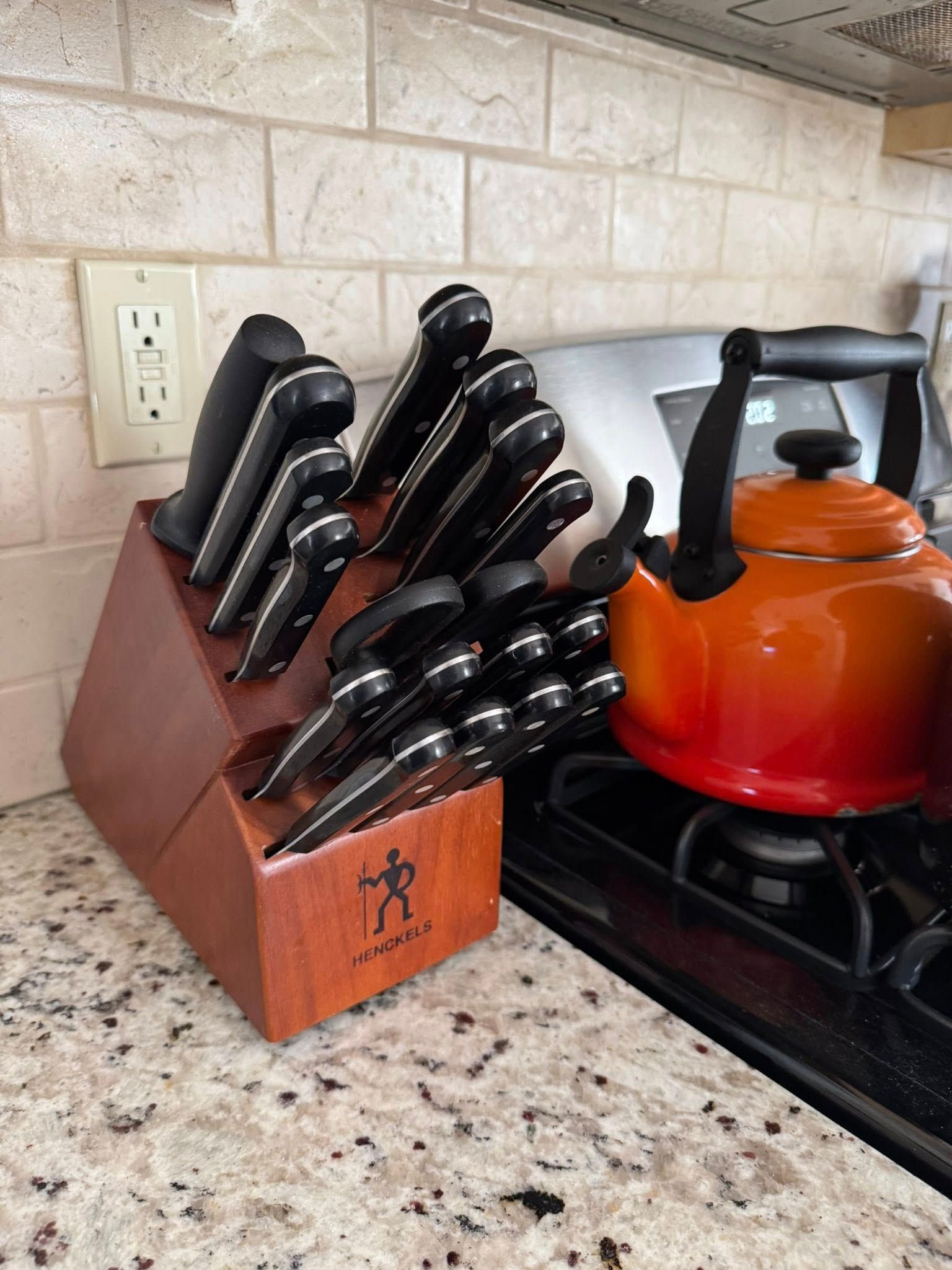 Knife block with various black-handled knives, next to an orange kettle on a stovetop in a kitchen.