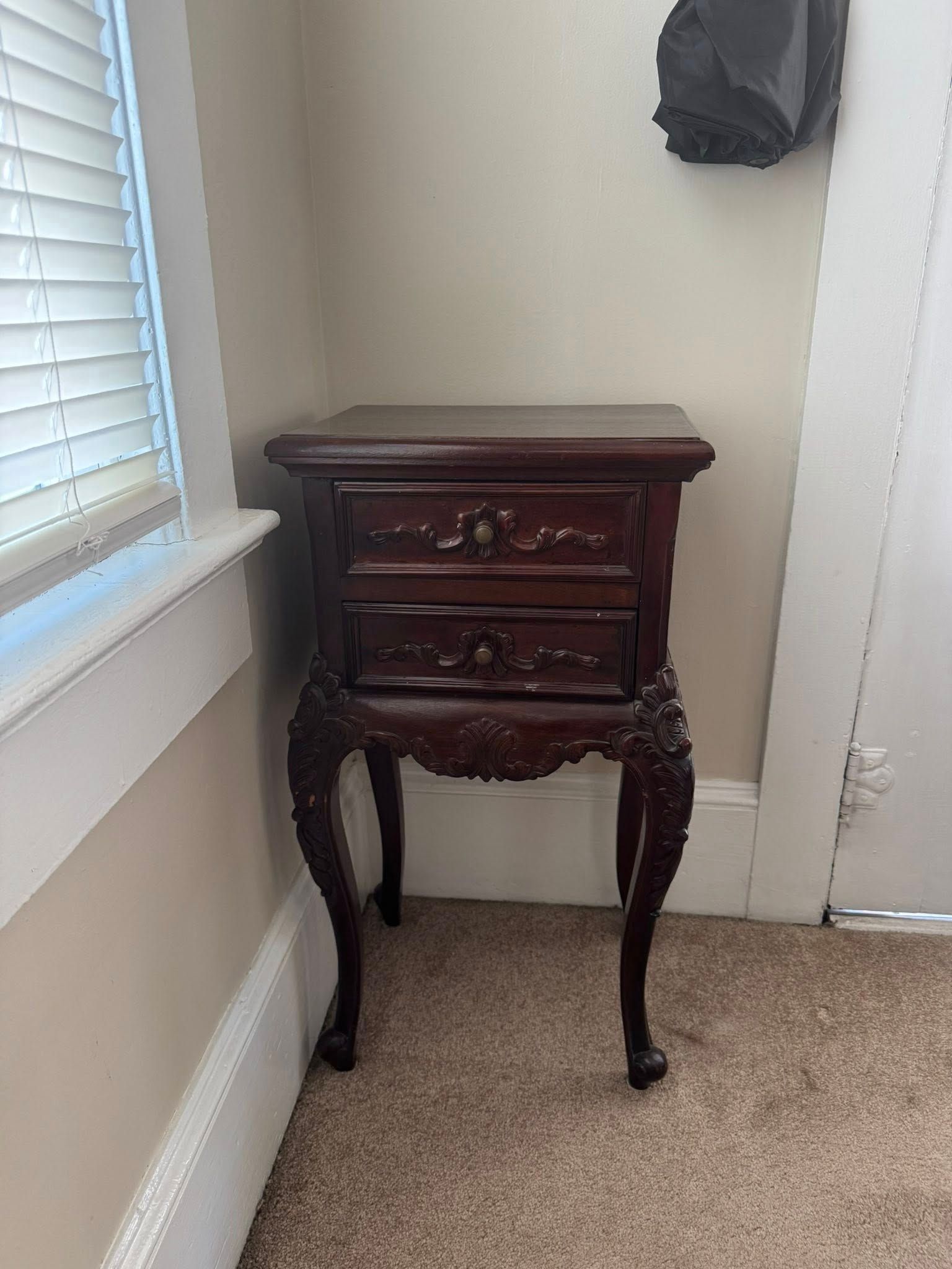 Dark wooden nightstand with two drawers and ornate legs, placed beside a window.