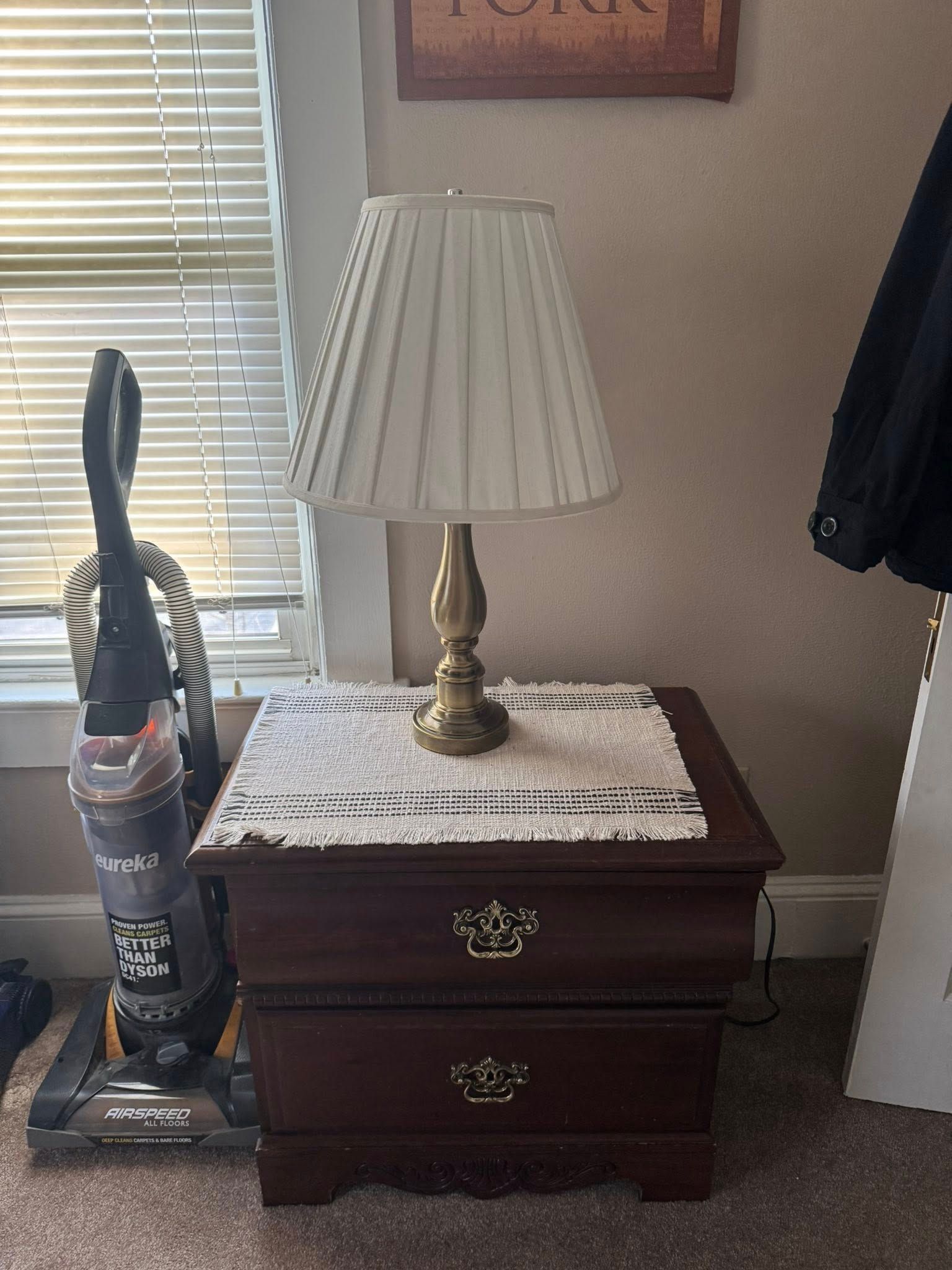 Nightstand with lamp, vacuum cleaner, and doily against a wall.