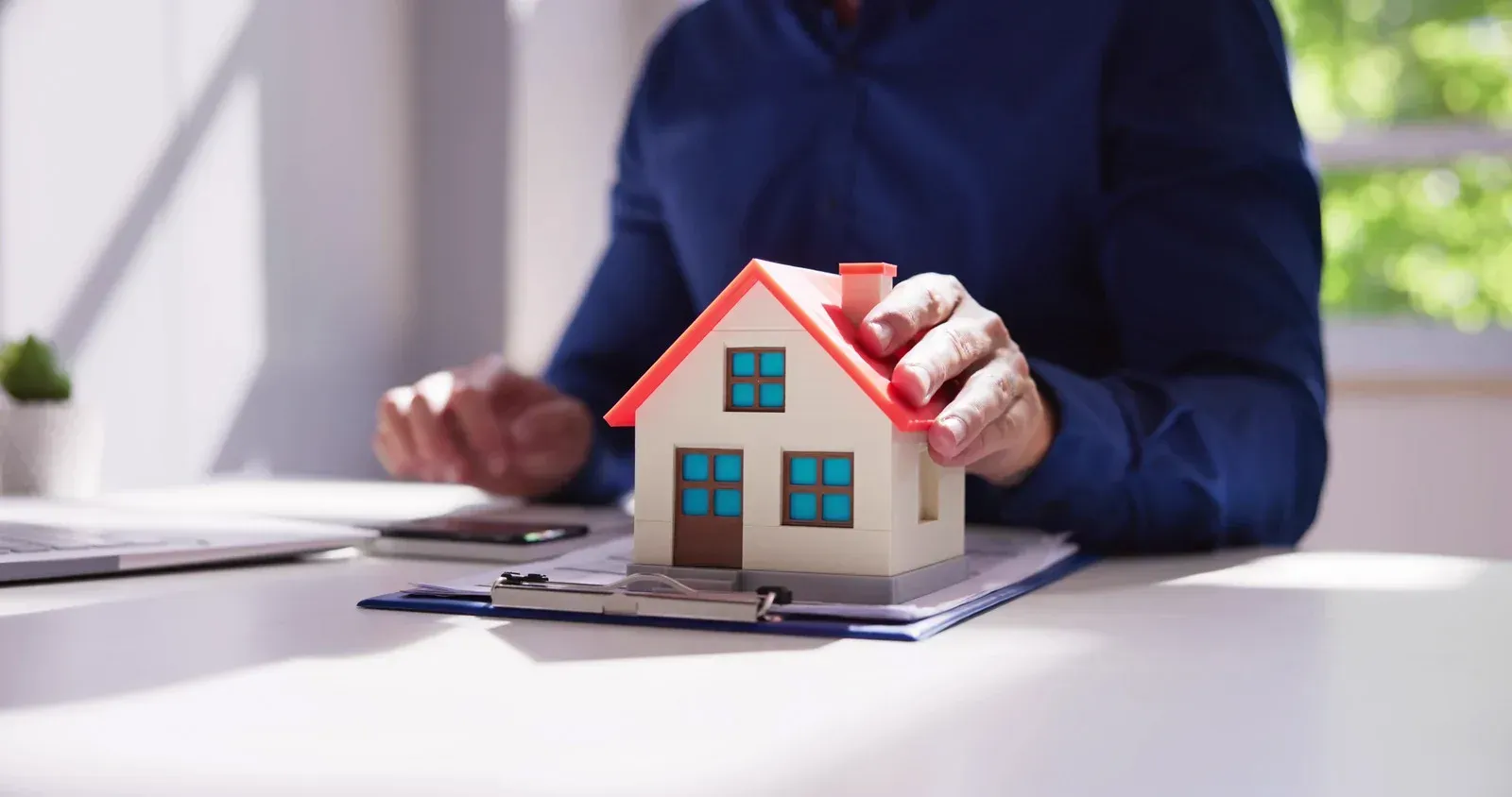 Person holding a model house over a clipboard and documents on a desk.