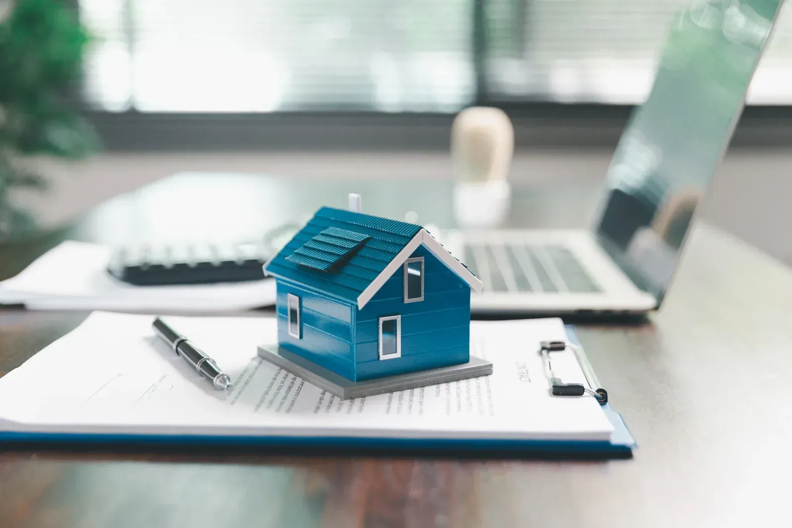 Person calculating with calculator, pointing at paperwork, model houses and red-roofed house on a desk.