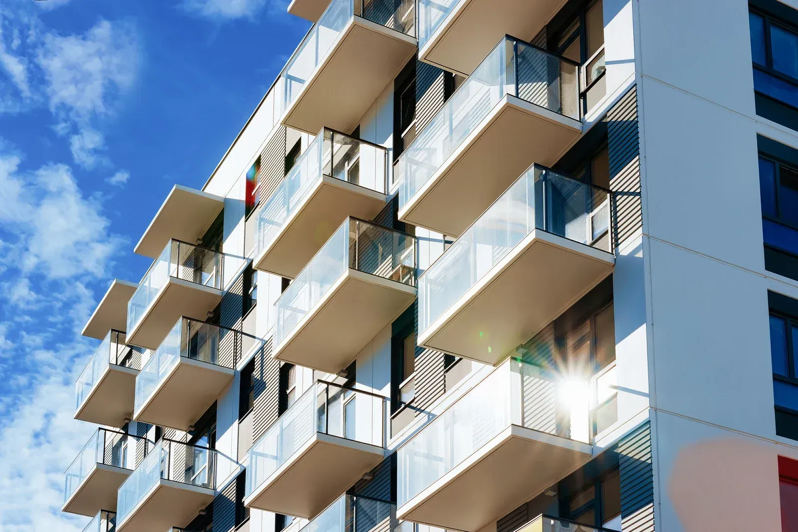 Modern apartment building with white facade and glass-railed balconies against a blue sky with sunlight.
