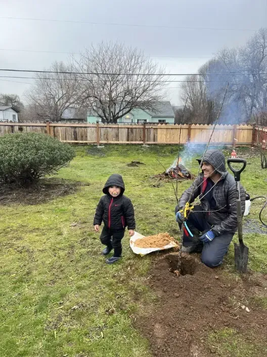 Child and person planting in backyard. Smoke from a fire visible. Overcast day.