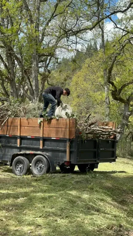 Man loading brush into a trailer in a grassy area with trees.