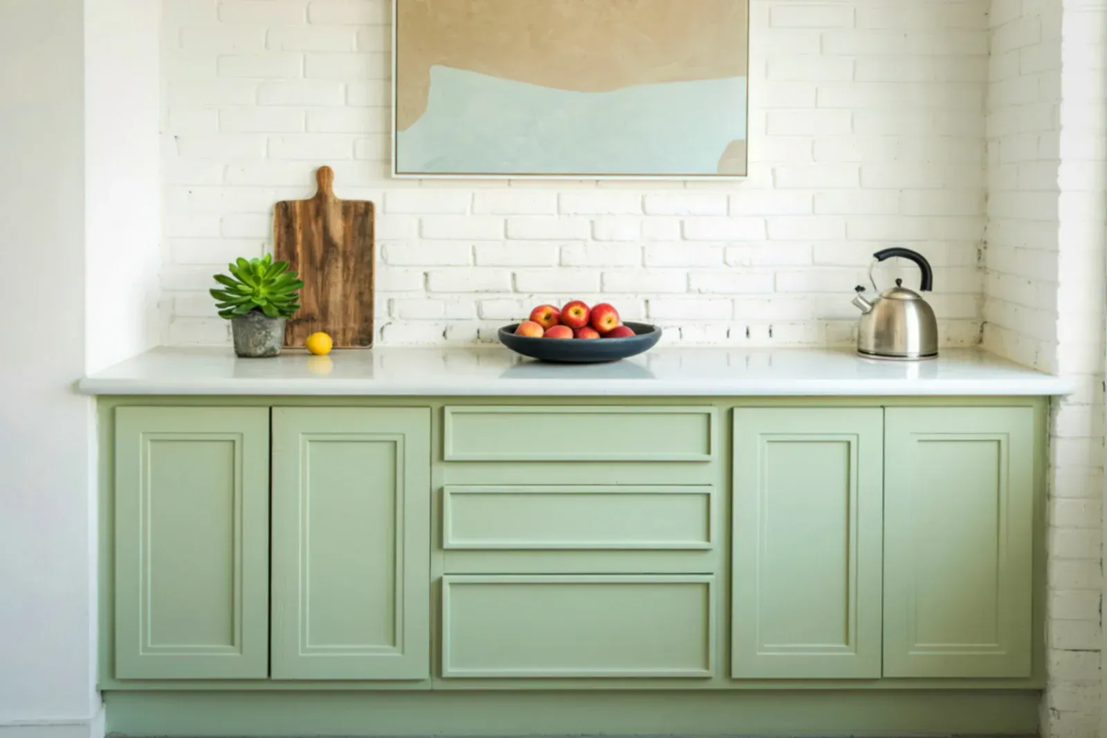 Green kitchen cabinets with white countertop, brick wall, fruit, cutting board, and art.