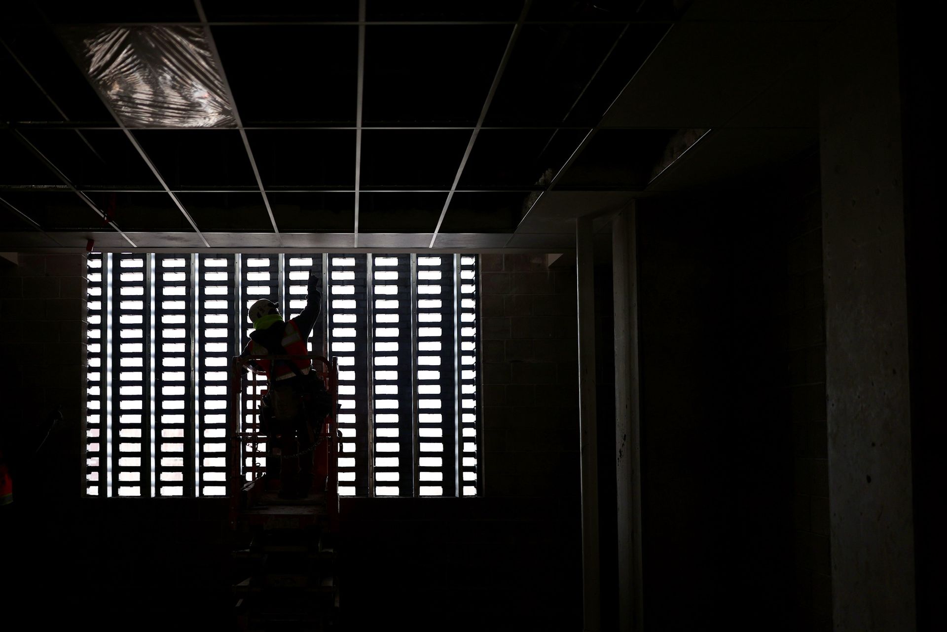 Construction worker silhouetted against a grid-patterned window, installing ceiling tiles in a dimly lit room on a Colorado construction site.