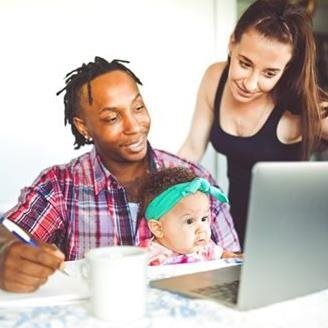 Man, baby, and woman looking at laptop. Man writes on paper. Baby looks at the laptop. Woman smiles.