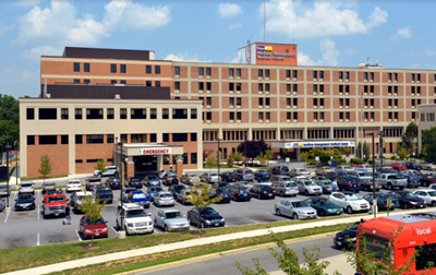Hospital building with emergency entrance, parking lot full of cars, blue sky.