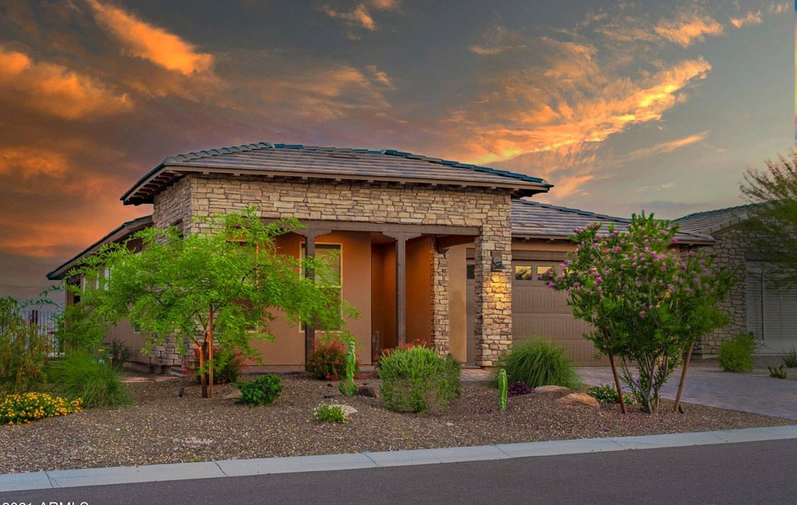 Stone-faced home with a tan exterior, dark roof, and lush landscaping against a sunset sky.
