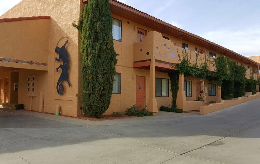 Two-story adobe building with a large lizard art on the wall, with a tall green tree on the side.