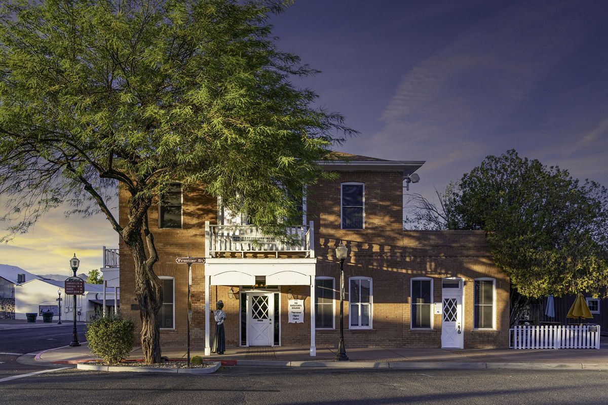 A two-story brown building with a white balcony and fence, shaded by a large tree under a dusk sky in a small town.