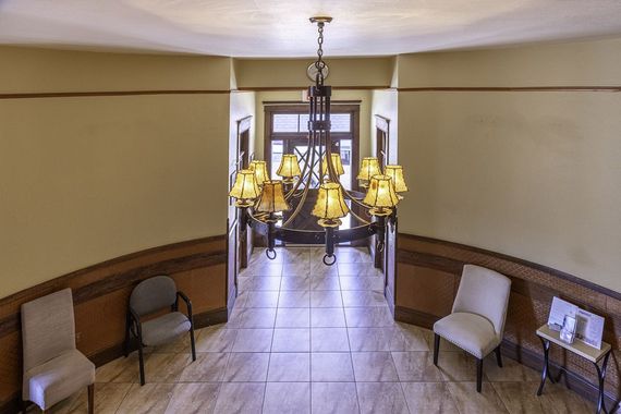 A beige, wood-paneled hallway with an ornate chandelier, tiled floors, three chairs, and a small side table.