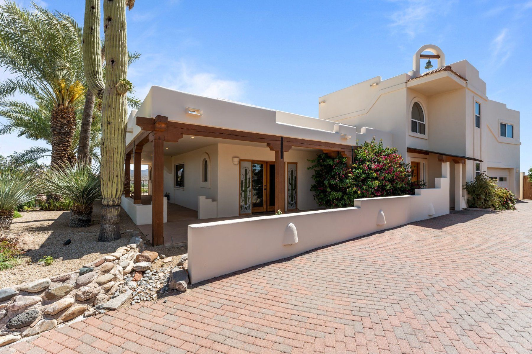 Spanish-style house with brick driveway, wooden beams, and palm trees against a blue sky.