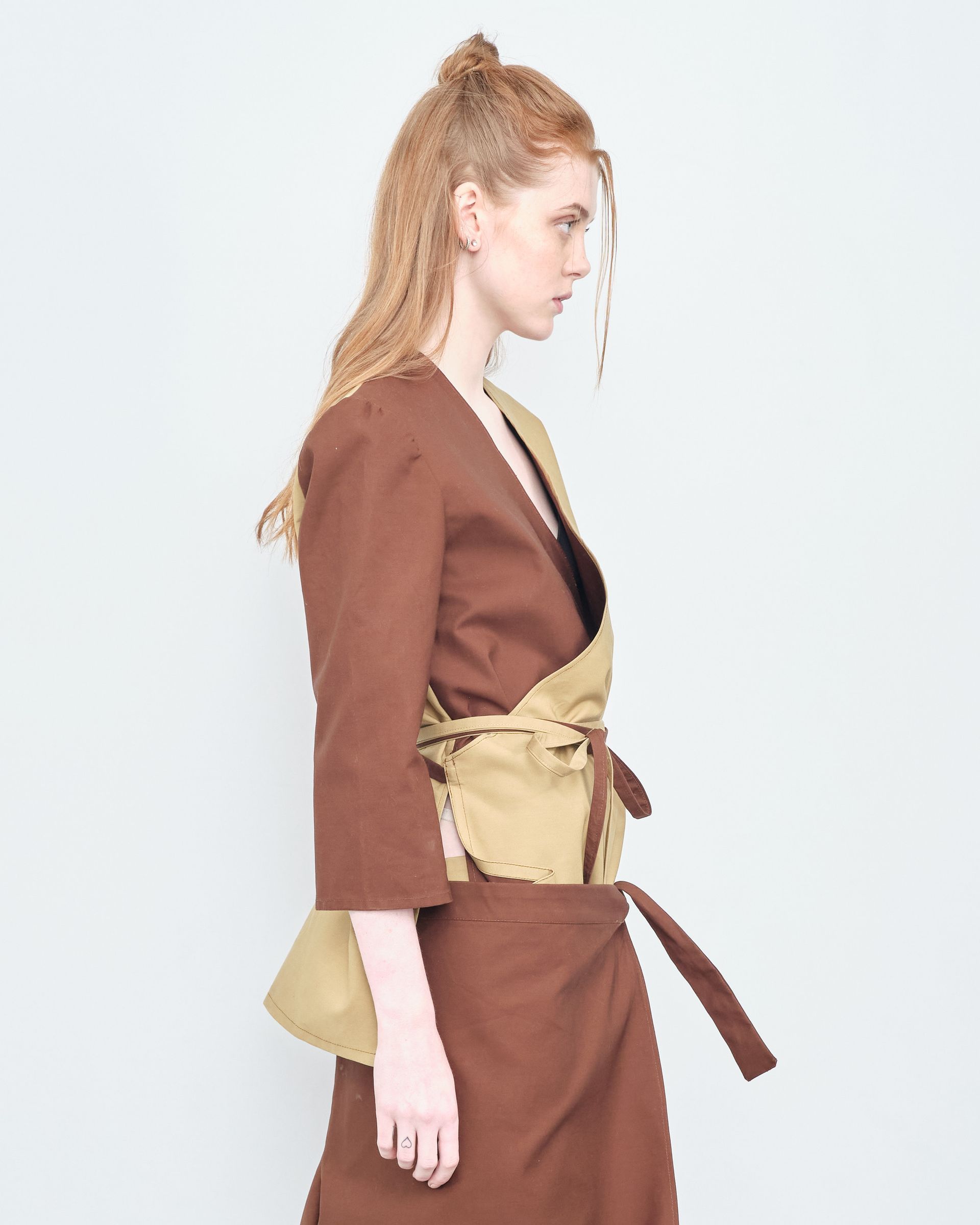 A woman wearing a brown jacket and a brown skirt is standing in front of a white wall.