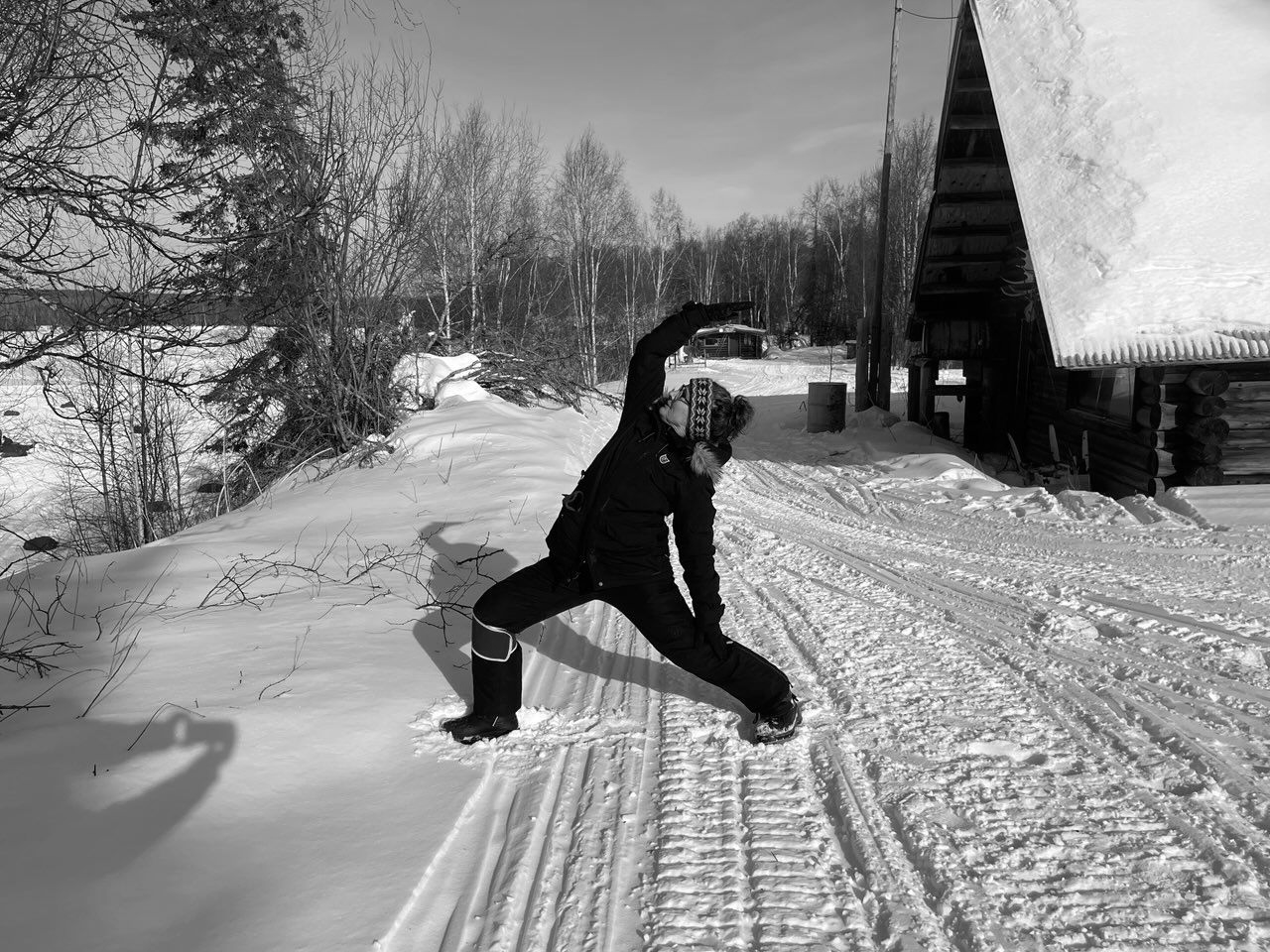 Person posing in a snowy yard near a cabin, with trees and a snow-covered path.