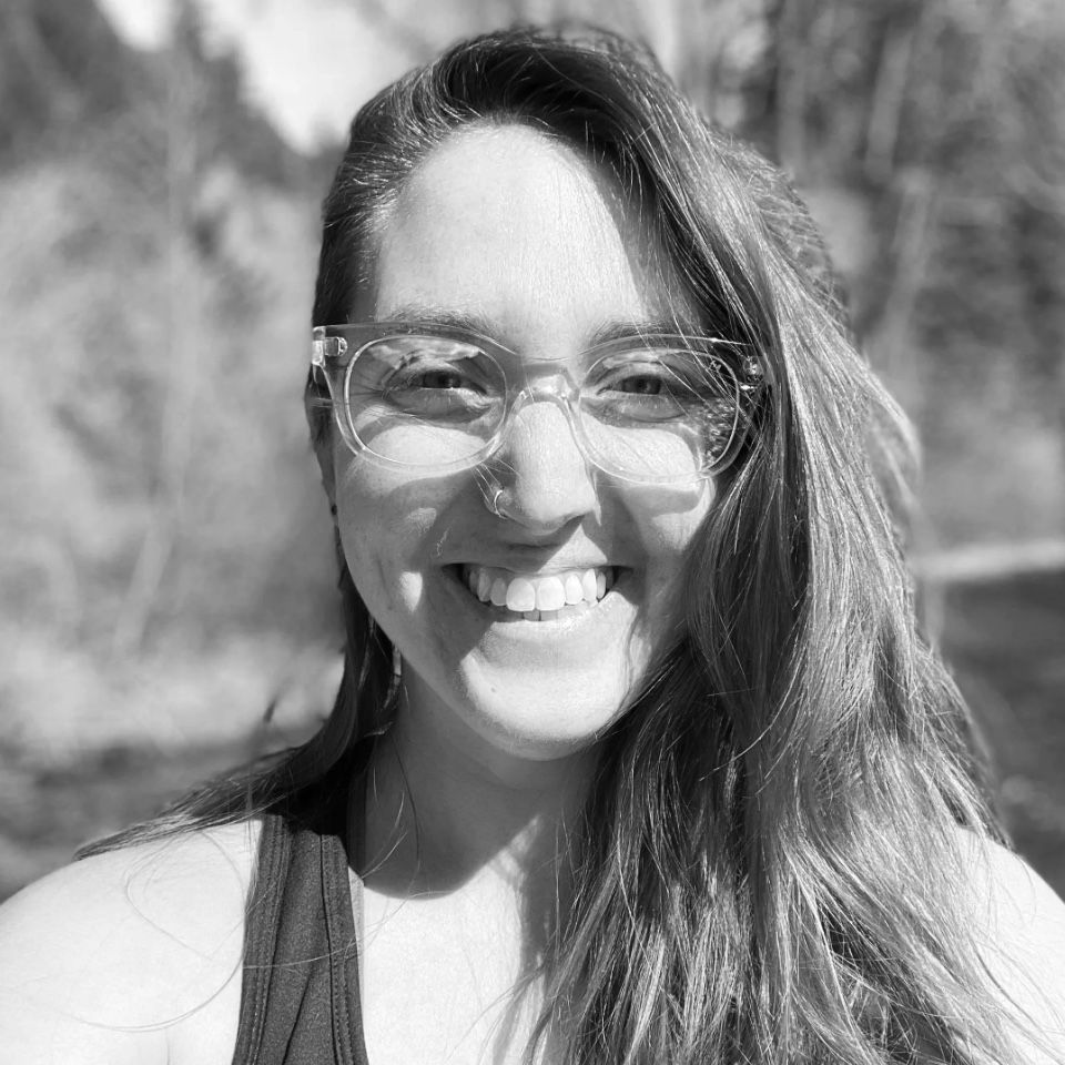 Smiling woman with glasses outdoors in bright sunlight, with trees blurred in the background