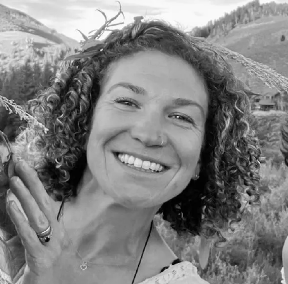 Smiling woman with curly hair outdoors in a mountain meadow, waving at the camera.