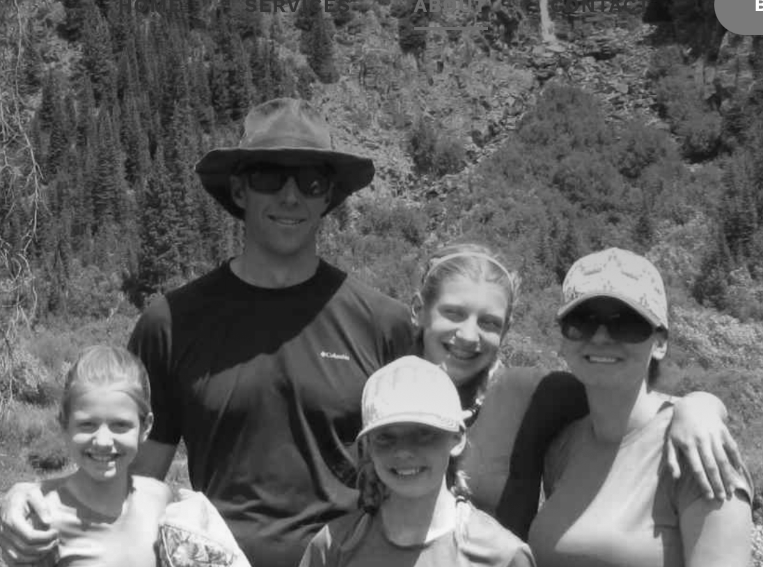 Five hikers smiling on a mountain trail, with trees and rocky cliffs in the background.