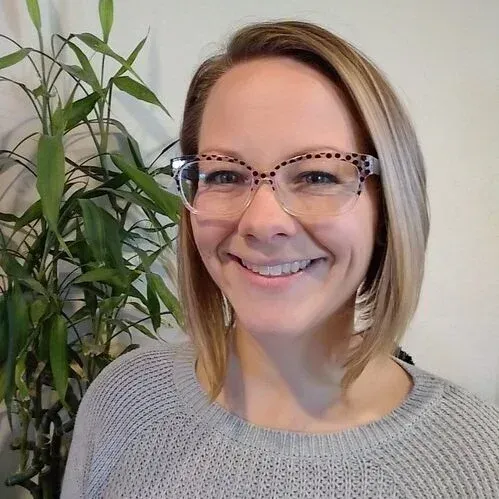 Smiling person with glasses in a gray sweater, standing beside a leafy houseplant.