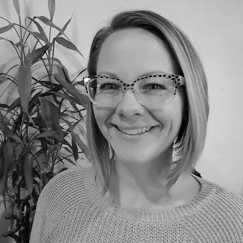 Smiling person with glasses standing beside a leafy plant in black and white.