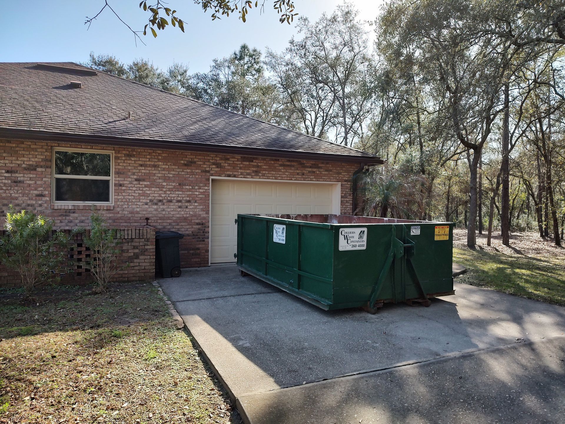 A green dumpster is parked in front of a brick house.